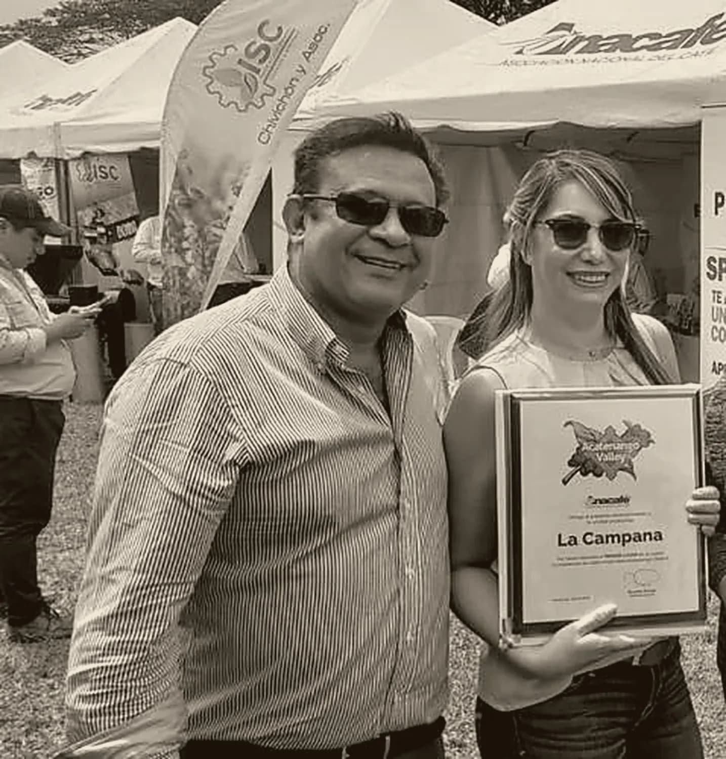 A man and a woman at an outdoor Anacafé event holding a diploma that reads “La Campana”.